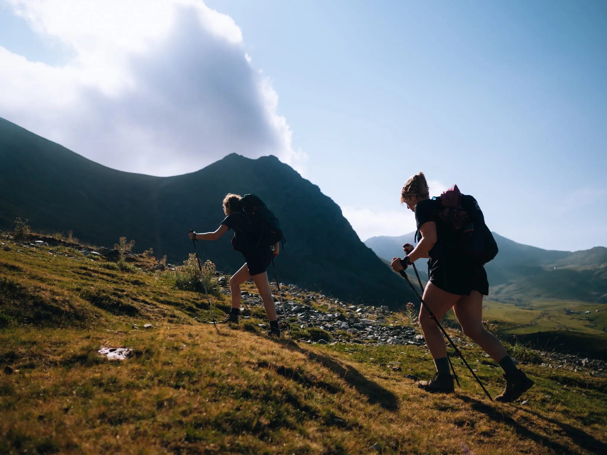 Zwei Wanderer mit Rucksäcken und Trekkingstöcken steigen einen sonnigen Grashang hinauf, dahinter eine Bergkette