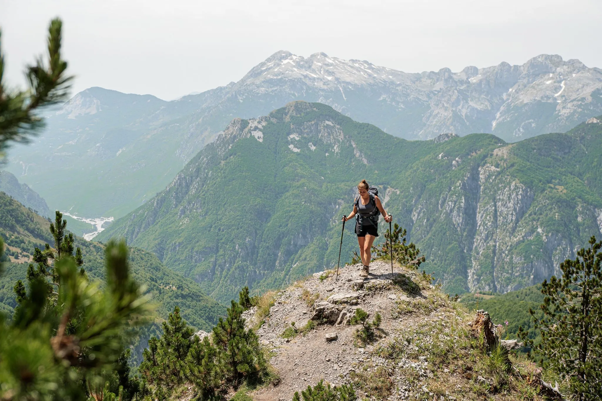Eine Frau mit Rucksack und Trekkingstöcken wandert über einen felsigen Grat, in der Ferne Gipfel mit Schneeresten