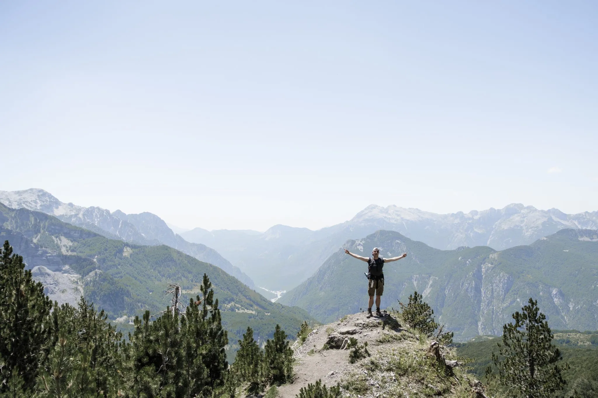 Ein Wanderer mit ausgebreiteten Armen auf einem felsigen Gipfel über einem weiten Tal in den Albanischen Alpen